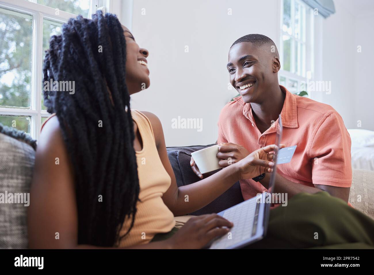 Glückliches junges afroamerikanisches Paar, das sich zu Hause unterhalten und sich anfreunden kann. Hübscher Mann, der Kaffee trinkt, während die Freundin online einkauft. Lächelnde Frau mit Kreditkarte und Laptop für die Bezahlung Stockfoto