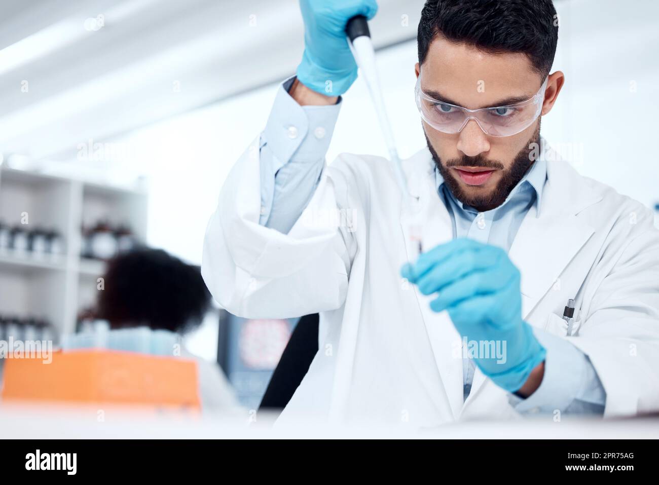 Ein männlicher Wissenschaftler gemischter Rassen trägt Schutzbrille und Labormantel, während er ein medizinisches Forschungsexperiment mit Pipette und Reagenzgläsern in einem Labor durchführt. Aufzeichnung seiner Ergebnisse für zukünftige Untersuchungen Stockfoto