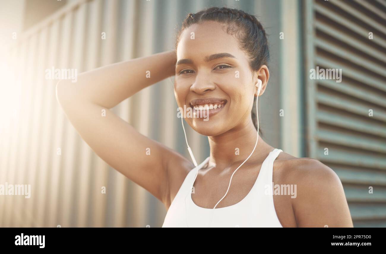 Porträt einer selbstbewussten jungen hispanischen Frau, die Musik mit Kopfhörern hört, während sie draußen in einer urbanen Umgebung trainiert. Entschlossene Sportlerin, die beim Training glücklich und motiviert aussieht Stockfoto