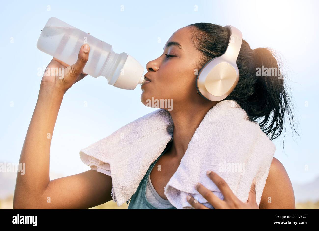 Eine Frau mit aktiver Passform, die nach dem Training im Freien kabellose Kopfhörer trägt und Wasser aus einer Flasche trinkt. Sportlerinnen löschen Durst und kühlen sich nach dem Training im Freien mit einem Handtuch ab Stockfoto