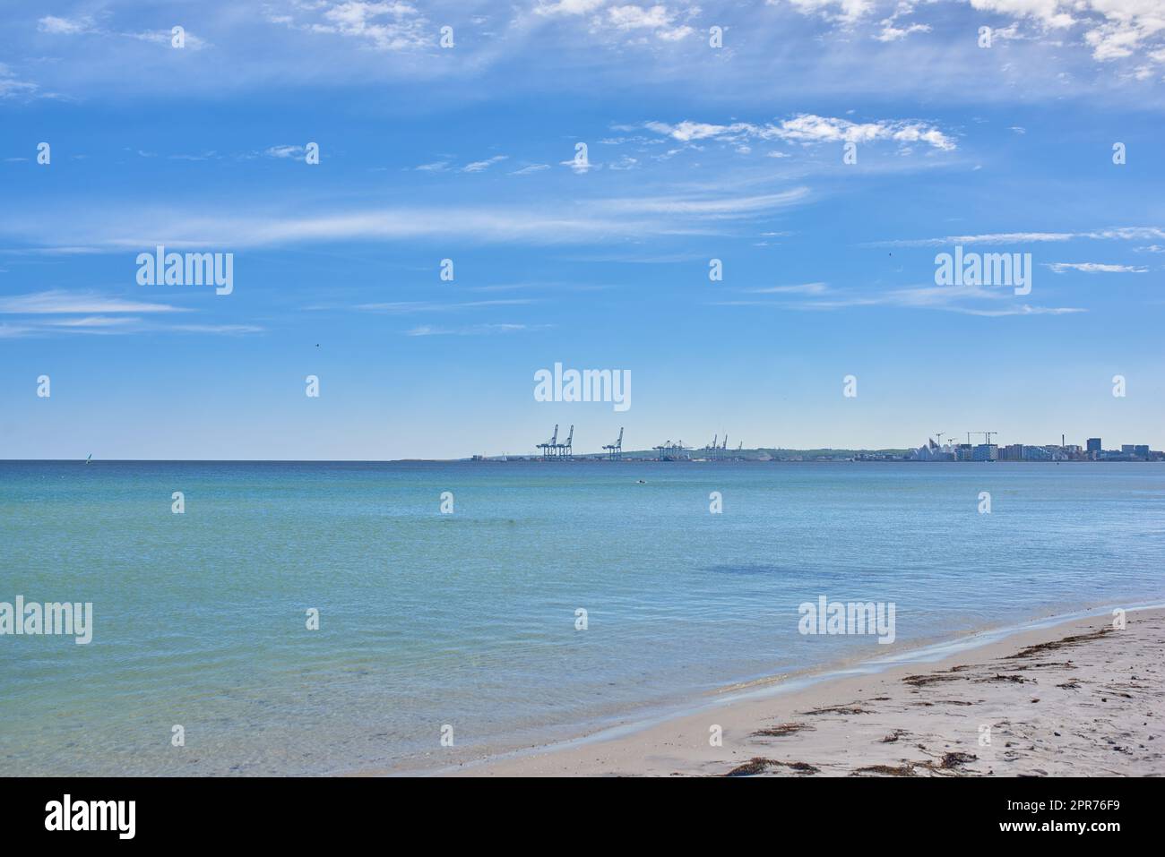 Kopiere den Raum am Meer mit blauem Hintergrund. Ruhige Meereswellen werden an einem leeren Strand mit einem Hafen am Horizont auf Steine gespült. Eine malerische Landschaft für einen entspannenden Sommerurlaub Stockfoto