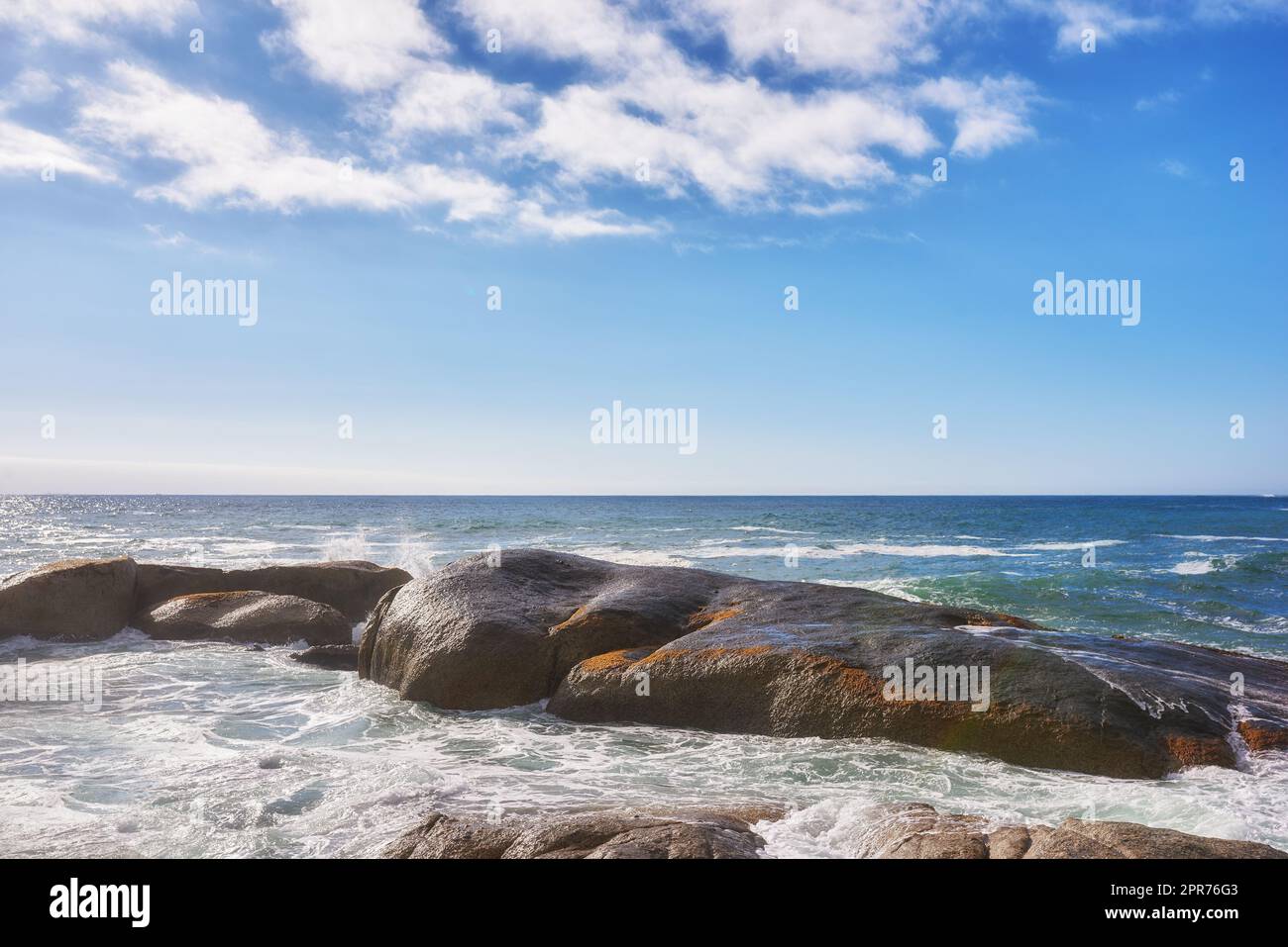 Landschaft mit großen Felsen im Ozean mit einem wolkigen blauen Himmel und Kopierraum. Am beliebten Camps Bay Beach in Kapstadt, Südafrika, spritzen Wellen gegen Felsen. Ein wunderschönes Sommerziel Stockfoto