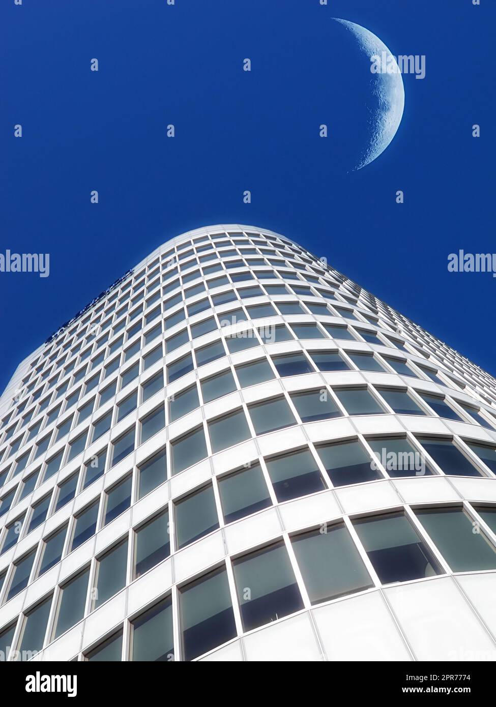 Ein halber Mond über einem Wolkenkratzer, der in hellblauem Himmel glänzt. Unter dem Halbmond des Viertelmondes über einer hohen modernen Strukturarchitektur, die dem untergebrachten Kapitalismus oder einer Wirtschaftskrise voraussagt Stockfoto