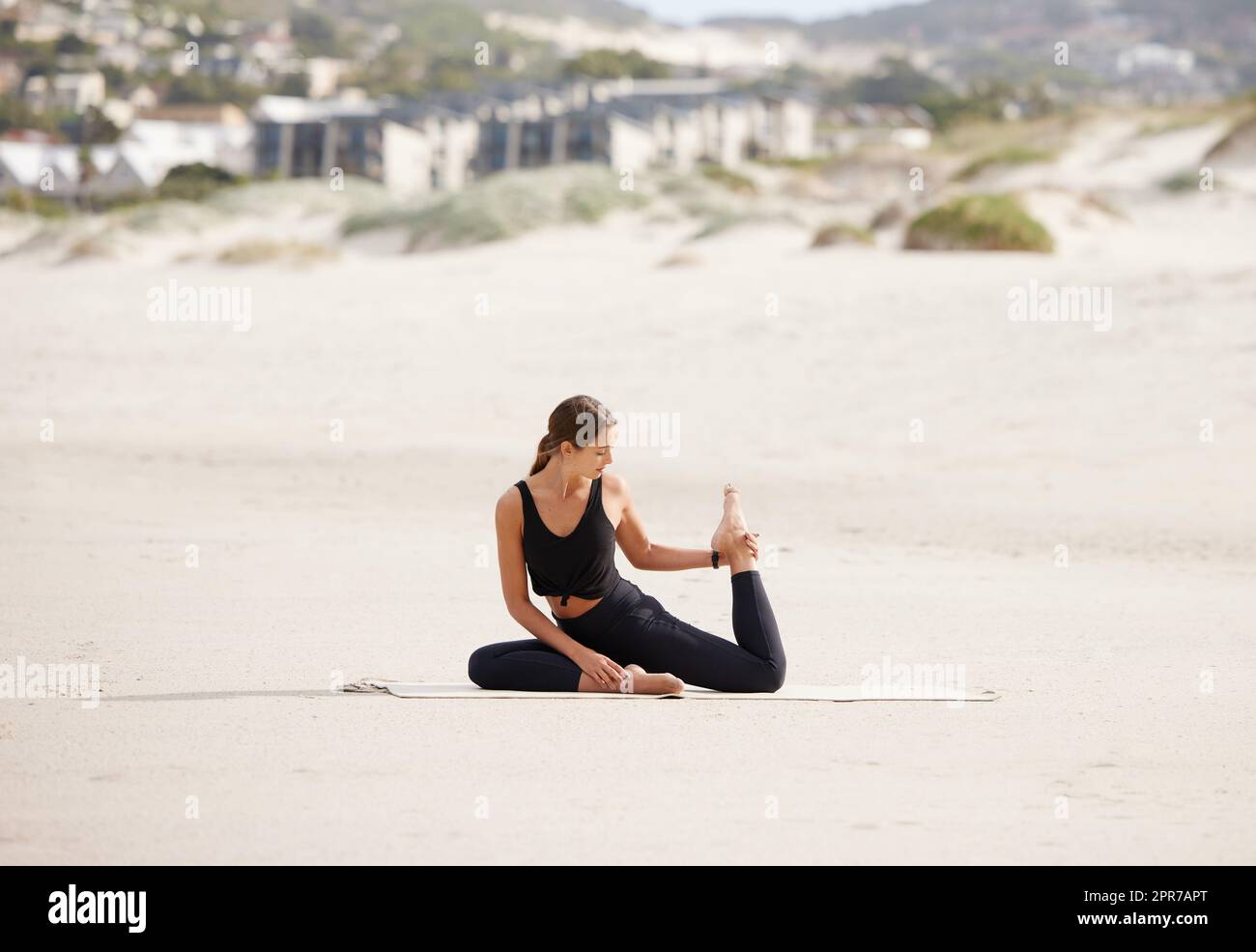 Das Meer bildet die perfekte Landschaft, um an sich selbst zu arbeiten. Eine junge Frau, die eine Taubenpose macht, während sie am Strand Yoga praktiziert. Stockfoto