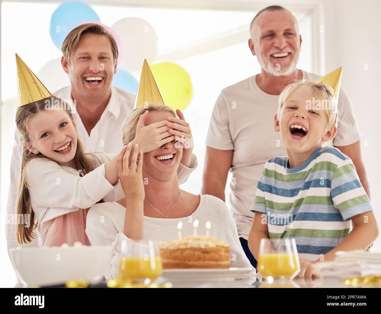 Kleines Mädchen, das der Mutter die Augen zudeckt, während es ihren Geburtstag feiert oder sie am Muttertag überrascht. Eine Frau feiert zu Hause mit ihrer Familie, trägt Partyhüte und amüsiert sich zusammen Stockfoto