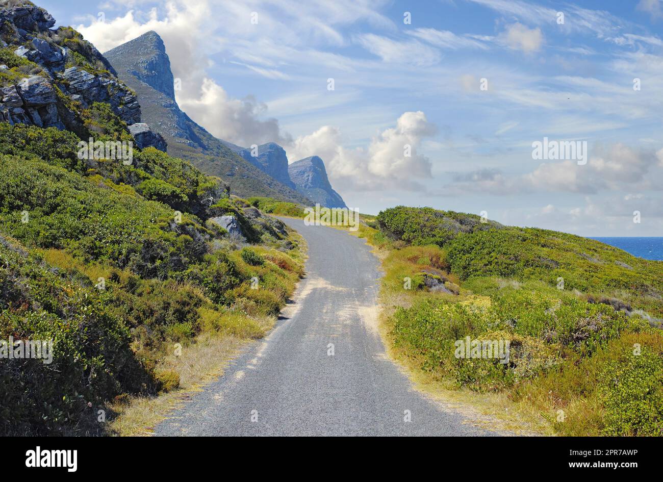 Asphaltstraße auf dem Tafelberg mit wolkenblauem Himmel. Landschaftslandschaft mit Landstraße für die Fahrt auf einem Bergpass entlang einer wunderschönen landschaftlich reizvollen Naturfahrt mit üppigen Sträuchern in Kapstadt, Südafrika Stockfoto