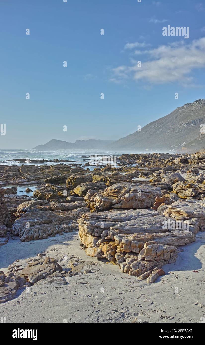 Copyspace am Meer mit blauem Himmel und felsiger Küste in Camps Bay, Kapstadt in Südafrika. Felsbrocken am Strand mit Tafelberg am Horizont an einem Sommertag Stockfoto