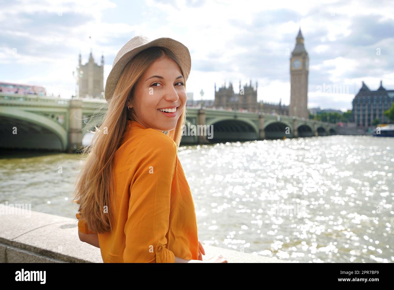 Schönes Mädchen in London, Großbritannien. Porträt einer jungen stilvollen Frau in orangefarbenem Hemd und Hut, die auf die Kamera blickt, mit Westminster Bridge im Hintergrund. Stockfoto