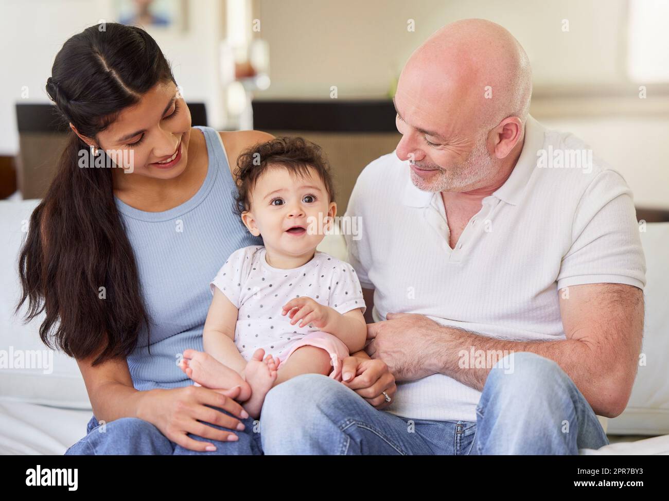 Ein junges, glückliches, gemischtrassiges Paar, das sich mit seinem adoptierten Baby zu Hause anfreundet. Ein kleines Mädchen lächelt, nachdem es von einem Weißen und einer Mischrasse adoptiert wurde. Mann und Frau entspannen sich mit ihrem Kind Stockfoto