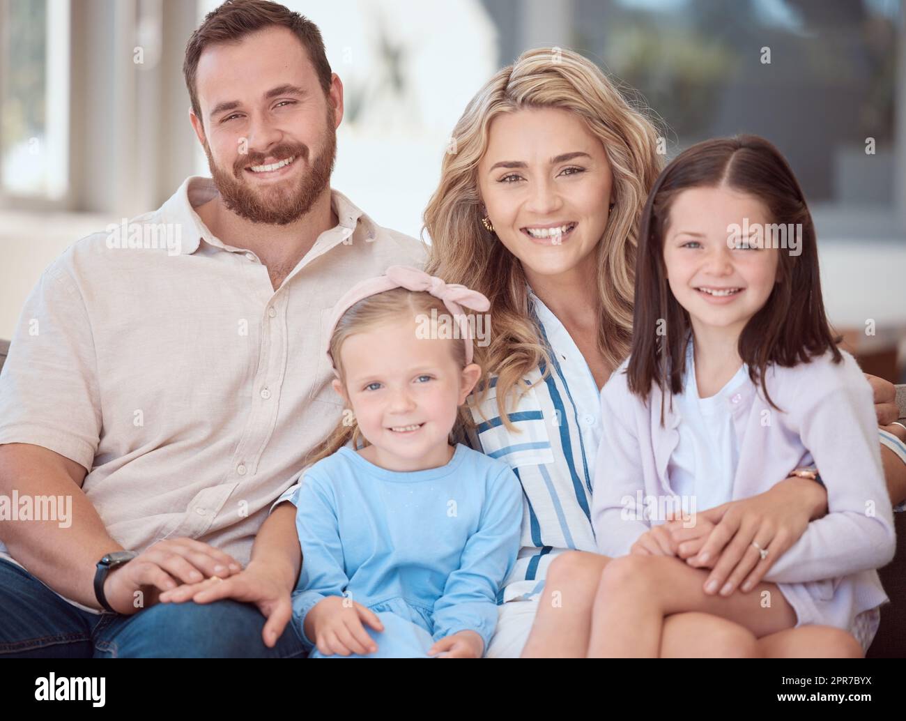 Porträt einer lächelnden jungen weißen Familie, die zu Hause auf dem Sofa dicht beieinander sitzt. Fröhliche, bezaubernde Mädchen, die sich an einem Wochenende mit ihren Eltern umarmen und eine Beziehung zu ihnen aufbauen. Glückliches Paar und Töchter Stockfoto