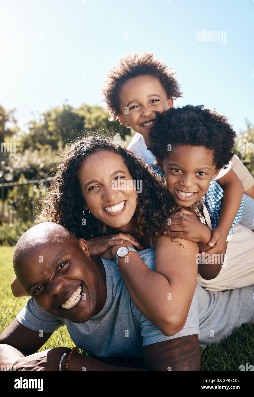 Eine glückliche afroamerikanische Familie von vier Personen, die übereinander liegen, während sie Spaß haben und zusammen in der Sonne spielen. Sorgenfreie Mutter und zwei Kinder stapelten sich auf Vater, während sie im Park eine Beziehung knüpften Stockfoto