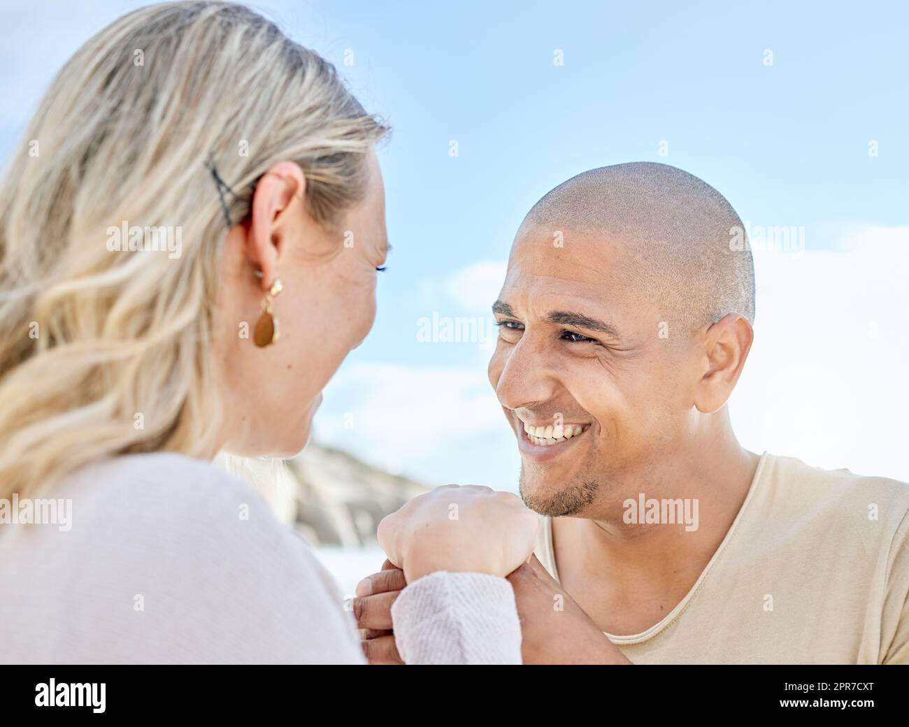Ein junger Mann, der seiner Freundin im Sommer am Strand einen Heiratsantrag macht. Ein glückliches gemischtrassiges Paar, das Händchen hält und lächelt. Bald Ehemann und Ehefrau, die draußen liebevoll in einen anderen Blick starren Stockfoto