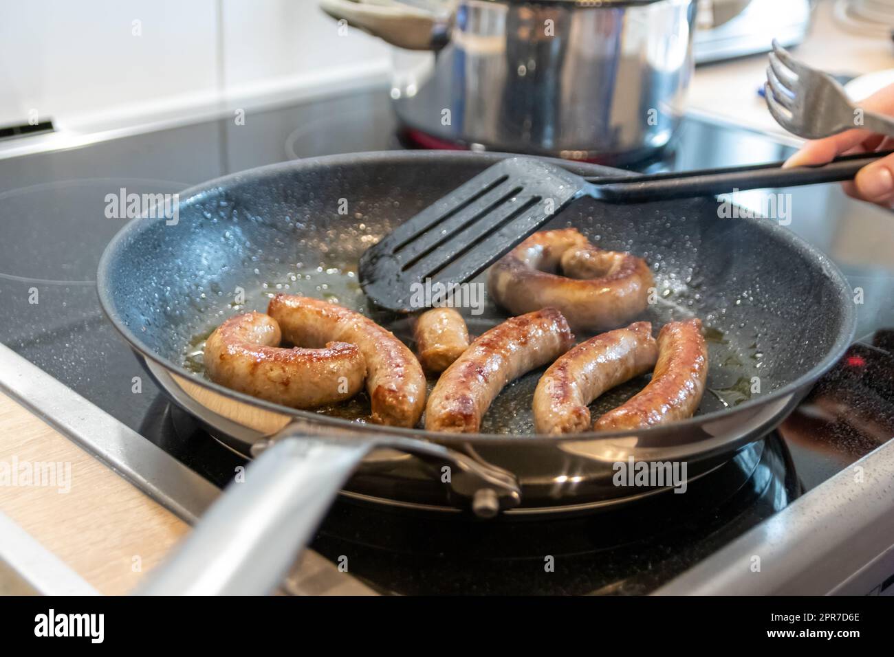 Frau, die Würstchen in der Pfanne auf Küchenplatte mit Gabel und schwarzem Spatel mit köstlicher fetter Bratwurst zum Abendessen kocht, als ungesunde, aber würzige und geröstete Wurstmahlzeit zum Mittagessen und die Kinder Stockfoto