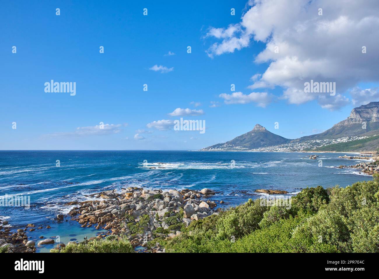 Eine Naturlandschaft mit einem ruhigen Meer und grünen Büschen, mit Lions Head Berg am Horizont, Kapstadt, Südafrika. Landschaft des Ozeans in der Nähe der Berge mit blauem Wolkenhimmel und Kopierraum. Stockfoto