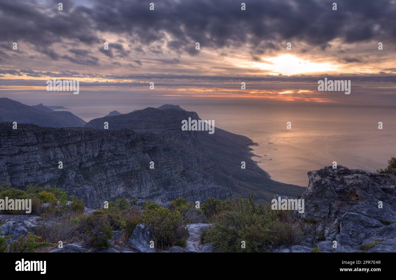 Blick über eine Bergküste bei Sonnenuntergang in Südafrika. Malerische Landschaft mit dunklen Wolken über einem ruhigen und friedlichen Ozean in der Nähe von Kapstadt mit der Sonne hinter grauen Wolken am Himmel und Kopierraum Stockfoto
