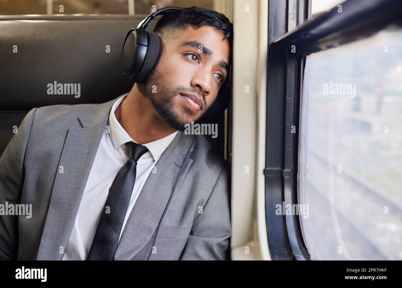 In tiefer Kontemplation während seines Arbeitsweges. Ein junger Geschäftsmann, der Kopfhörer trägt, während er während seines Arbeitsweges aus dem Fenster auf einen Zug starrt. Stockfoto