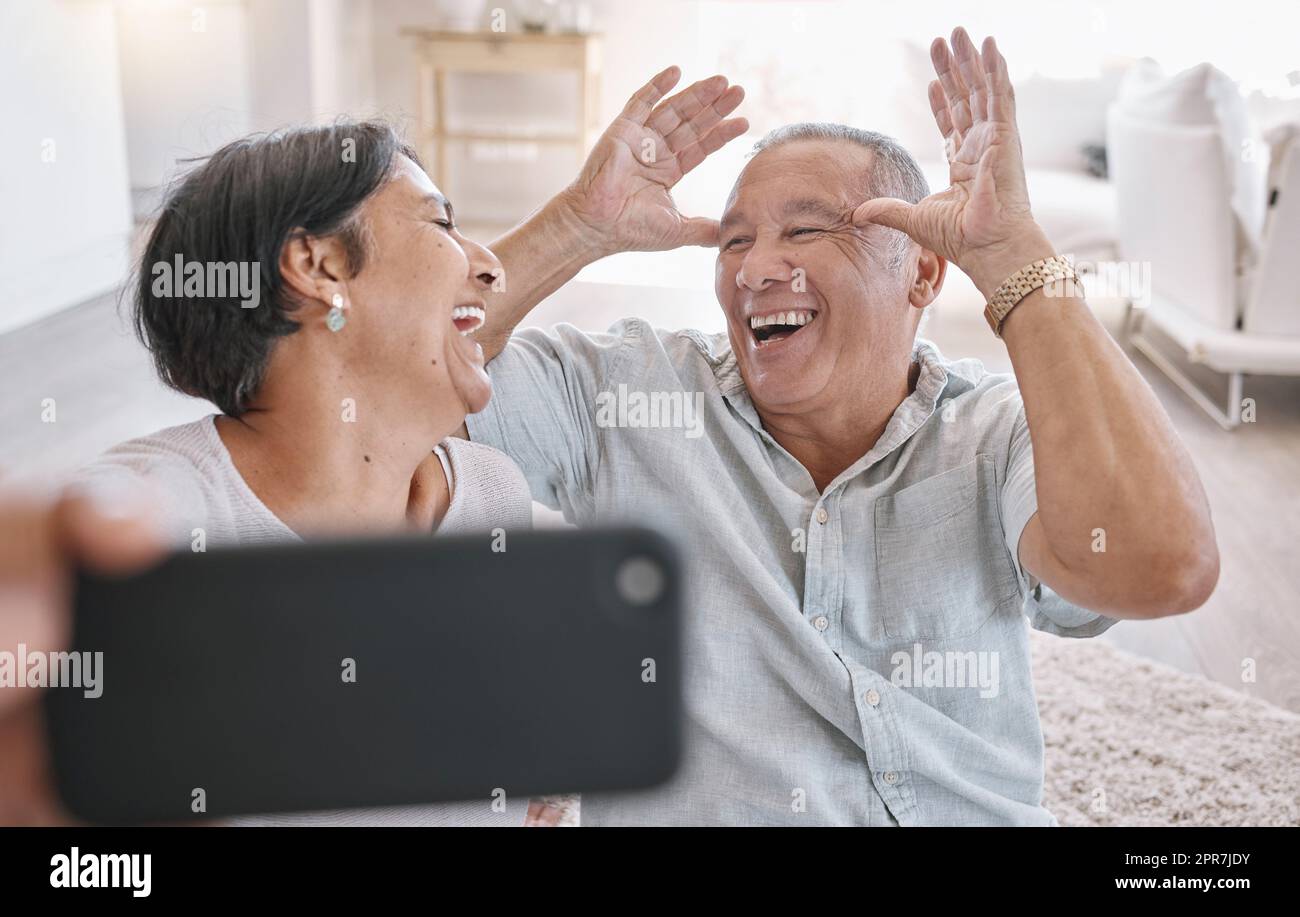 Nahaufnahme eines liebevollen Seniorenpaares, das Selfies macht, während es sich zu Hause im Wohnzimmer entspannt. Gemischtes Paar mit Spaß beim Fotografieren in der Lounge zu Hause Stockfoto