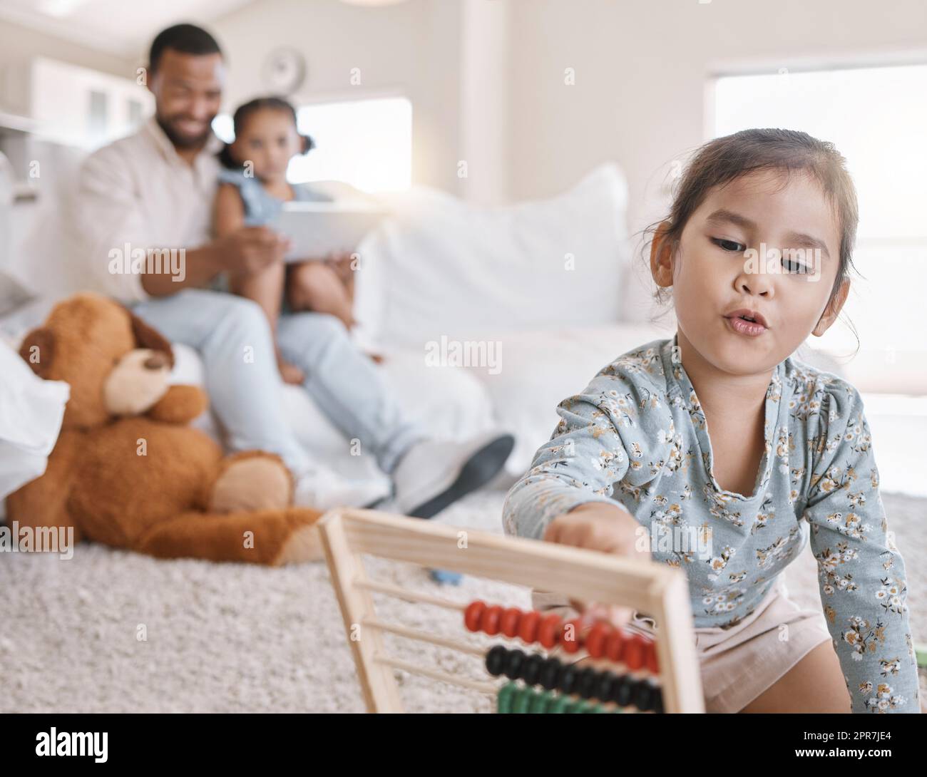 Nahaufnahme eines kleinen Mädchens mit Abakus, während sie im Wohnzimmer auf dem Boden saß, während ihr Vater und ihre Schwester zu Hause ein digitales Tablet auf dem Sofa benutzten Stockfoto
