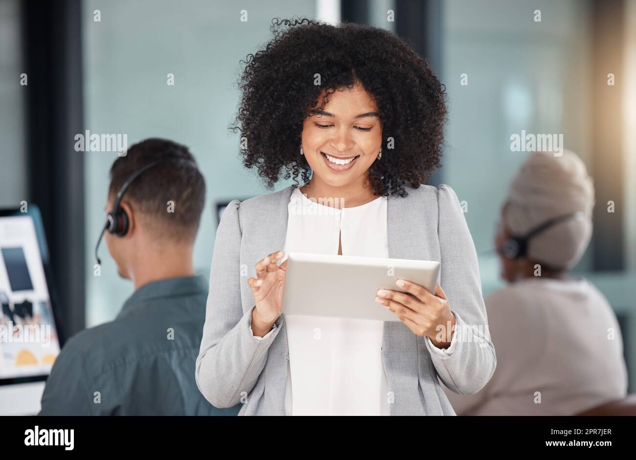 Junge Geschäftsfrau gemischter Rassen, die auf einem digitalen Tablet surft, während sie mit ihren Kollegen im Hintergrund in einem Callcenter arbeitet. Zufriedene Manager und Vorgesetzte planen online mit intelligenten Apps für den Kundendienst Stockfoto
