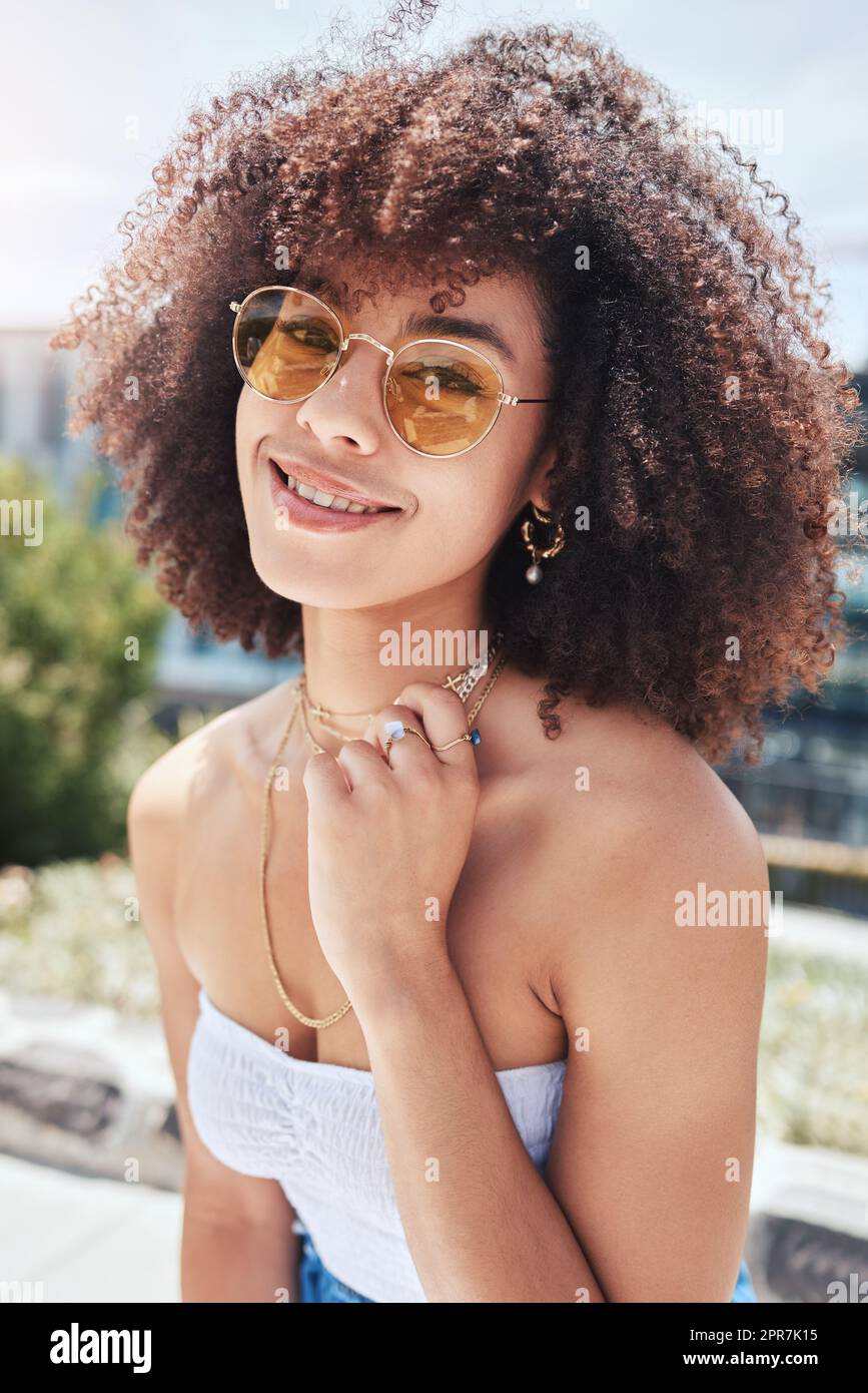 Porträt einer jungen, trendigen, wunderschönen Frau mit gemischter Rasse mit einem Afro, das lächelt und alleine draußen posiert. Eine hispanische Frau, die eine Sonnenbrille trägt und glücklich ist. Modische afroamerikanische Frau in der Stadt Stockfoto