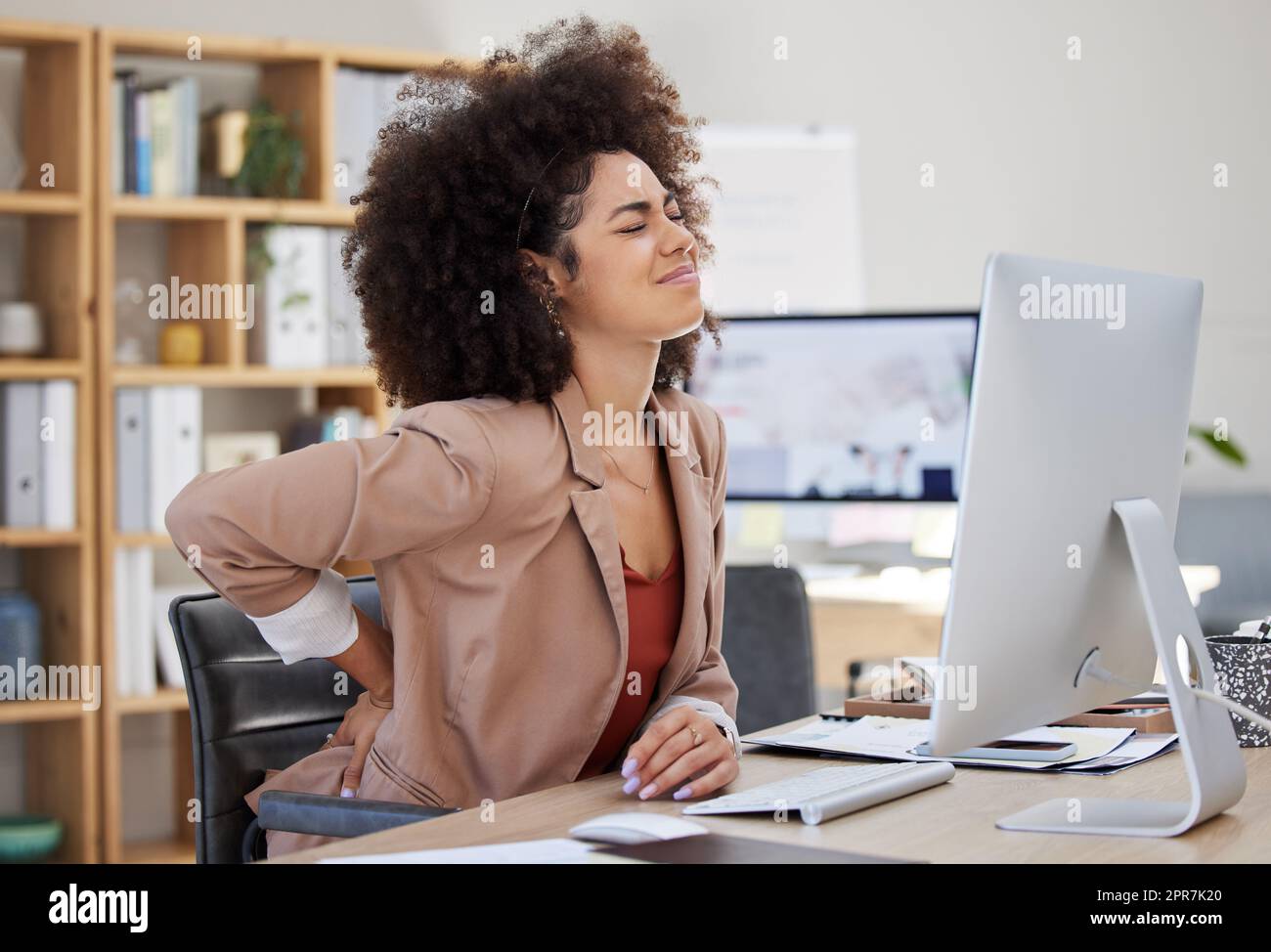 Eine junge Geschäftsfrau gemischter Rassen mit einem Afro, die Rückenschmerzen hat, während sie im Büro an ihrem Schreibtisch sitzt. Ein wunderschöner afroamerikanischer Unternehmer, der mit Krämpfen und Schmerzen kämpft Stockfoto