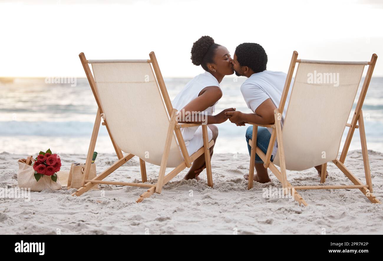 Ein glückliches afroamerikanisches Paar, das einen Tag am Meer verbringt. Inhalte, die Freund und Freundin küssen, während sie am Strand sitzen. Liebevoller Mann und Frau, die sich am Meer anfreunden Stockfoto