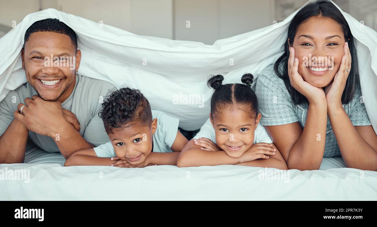 Porträt einer glücklichen Familie mit zwei Kindern, die unter der Bettdecke lagen und lächelnd vor die Kamera schauten. Ein Mädchen und ein Junge liegen im Bett mit ihren Eltern, die sich anfreunden und Spaß haben Stockfoto