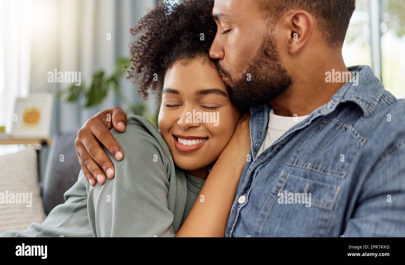 Ein junger, glücklicher Freund gemischter Rassen küsst seine Freundin auf die Stirn, während er sich zu Hause entspannt. Gemütlicher hispanischer Ehemann und Ehefrau, die lächeln, während sie sich zu Hause in der Lounge umarmen Stockfoto