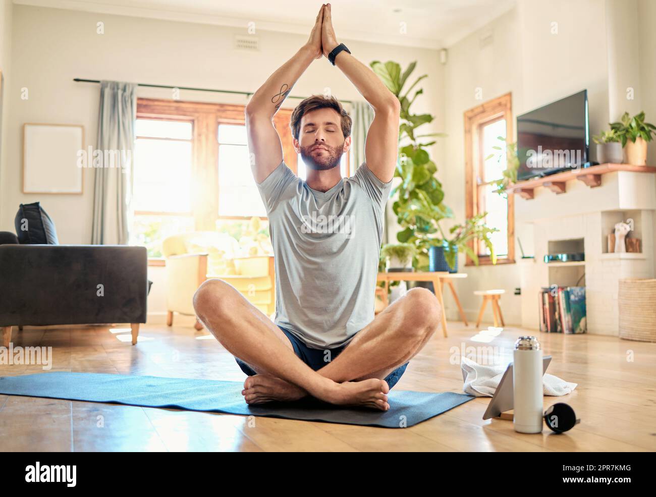 Ein junger weißer Mann sitzt mit gekreuzten Beinen und geschlossenen Augen und meditiert in Harmonie mit den Händen in namensvoller Geste, während er zu Hause Yoga praktiziert. Ein ruhiger, entspannter und fokussierter Kerl fühlt sich wie ein Zen und betet leise für Stressabbau und Frieden Stockfoto