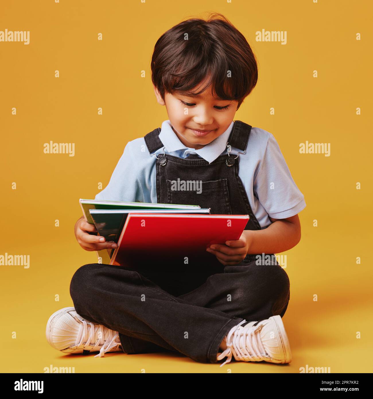 Ein süßer kleiner asiatischer Junge, der auf dem Boden sitzt und lässige Kleidung trägt, während er vor einem orangefarbenen Hintergrund liest. Glücklich und zufrieden, während der Schwerpunkt auf Bildung liegt. Das Kind ist bereit für die Schule Stockfoto
