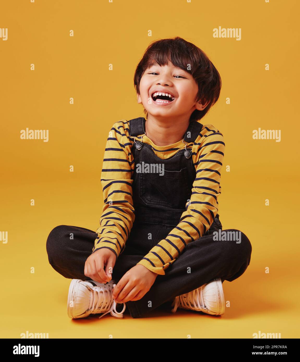 Ein süßer kleiner asiatischer Junge, der mit lässiger Kleidung auf dem Boden sitzt, während er lacht und seine Beine vor einem orangefarbenen Hintergrund kreuzt. Süßer, glücklicher kleiner Junge, sicher und allein Stockfoto