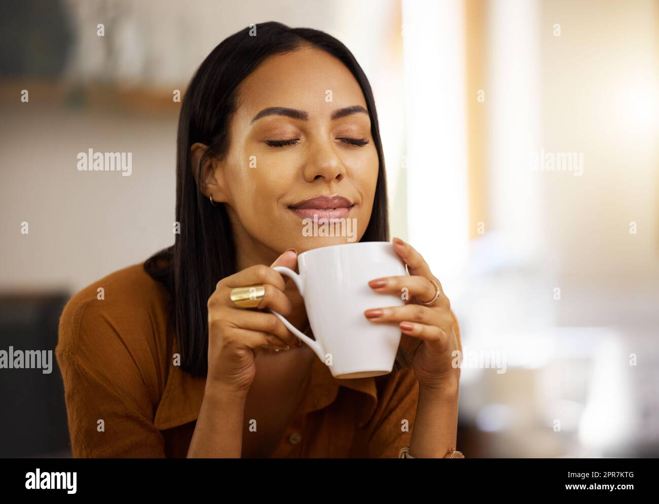 Junge, glückliche, wunderschöne Mischrasse Frau, die eine Tasse Kaffee allein zu Hause genießt. Hispanische Frau in ihren 20s, die lächelt, während sie eine Tasse Tee in der Küche zu Hause trinkt Stockfoto