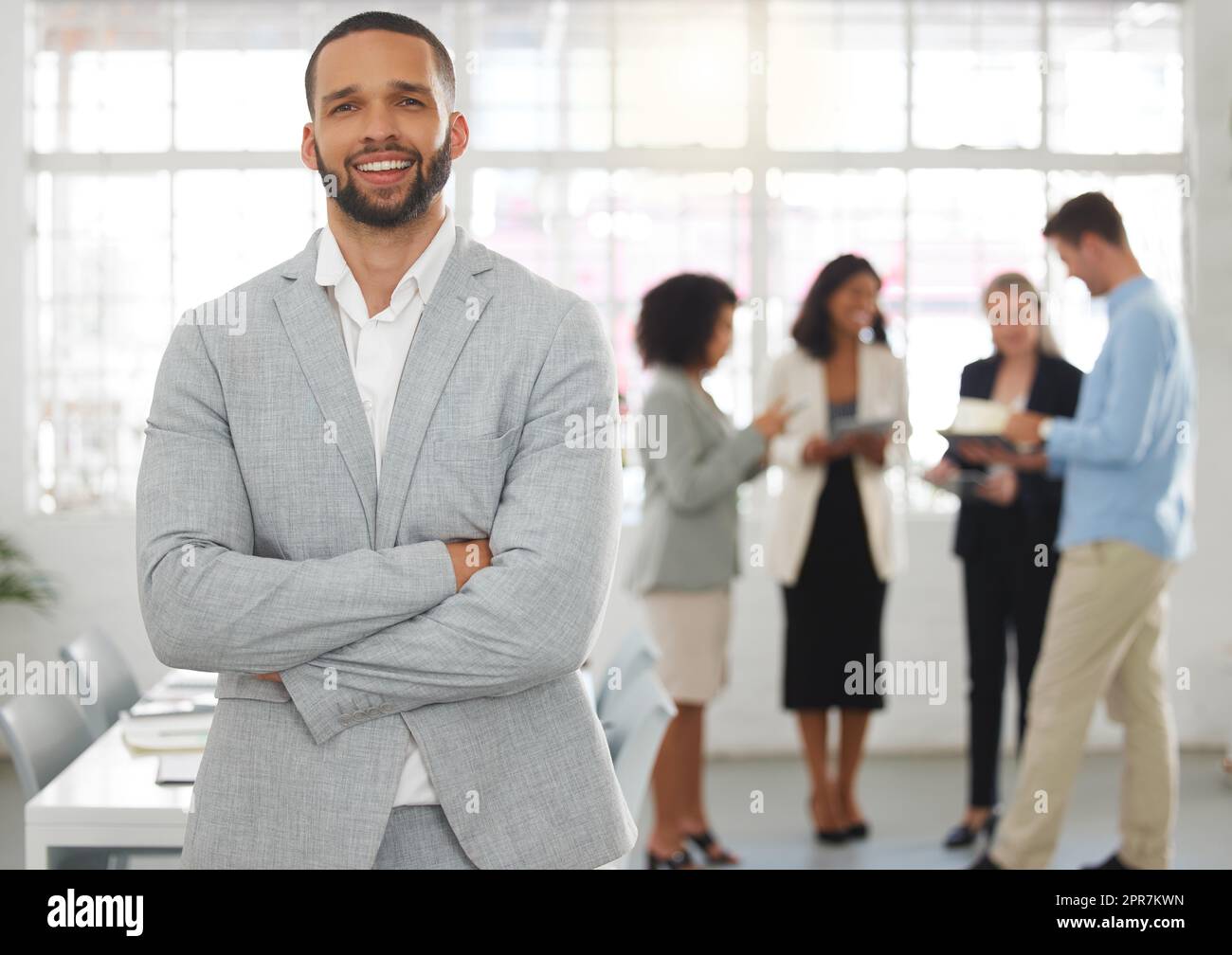 Junger glücklicher gemischter Geschäftsmann, der mit gekreuzten Armen in einem Büro mit Kollegen steht. Hispanischer männlicher Chef bei einem Treffen mit Kollegen Stockfoto