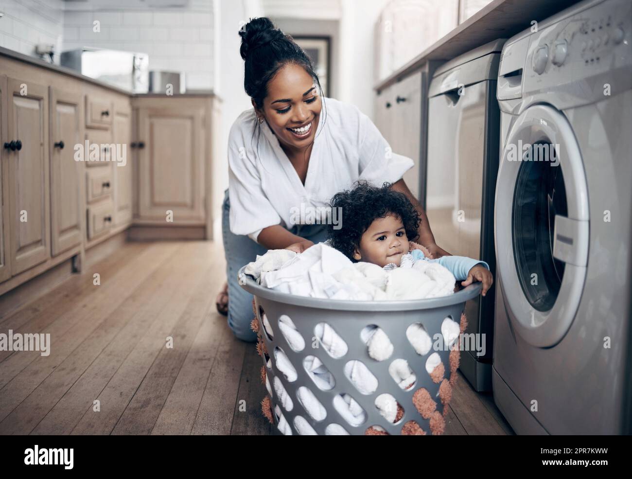 Eine junge Mutter, die sich spielerisch mit ihrem kleinen Mädchen verklebt, während sie die Wäsche zu Hause macht. Stockfoto