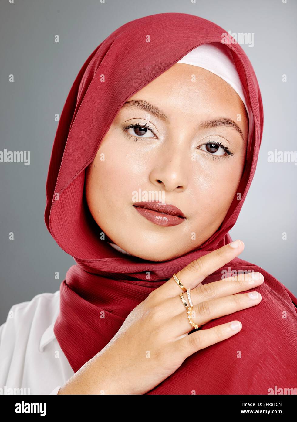 Porträt einer stilvollen muslimischen Frau mit Make-up und goldenen Ringen isoliert vor grauem Hintergrund. Die trendige junge Hijab-Dame fühlt sich sicher in ihren roten Schal und trendigen Kostümschmuck. Bescheidenheit und Schönheit Stockfoto