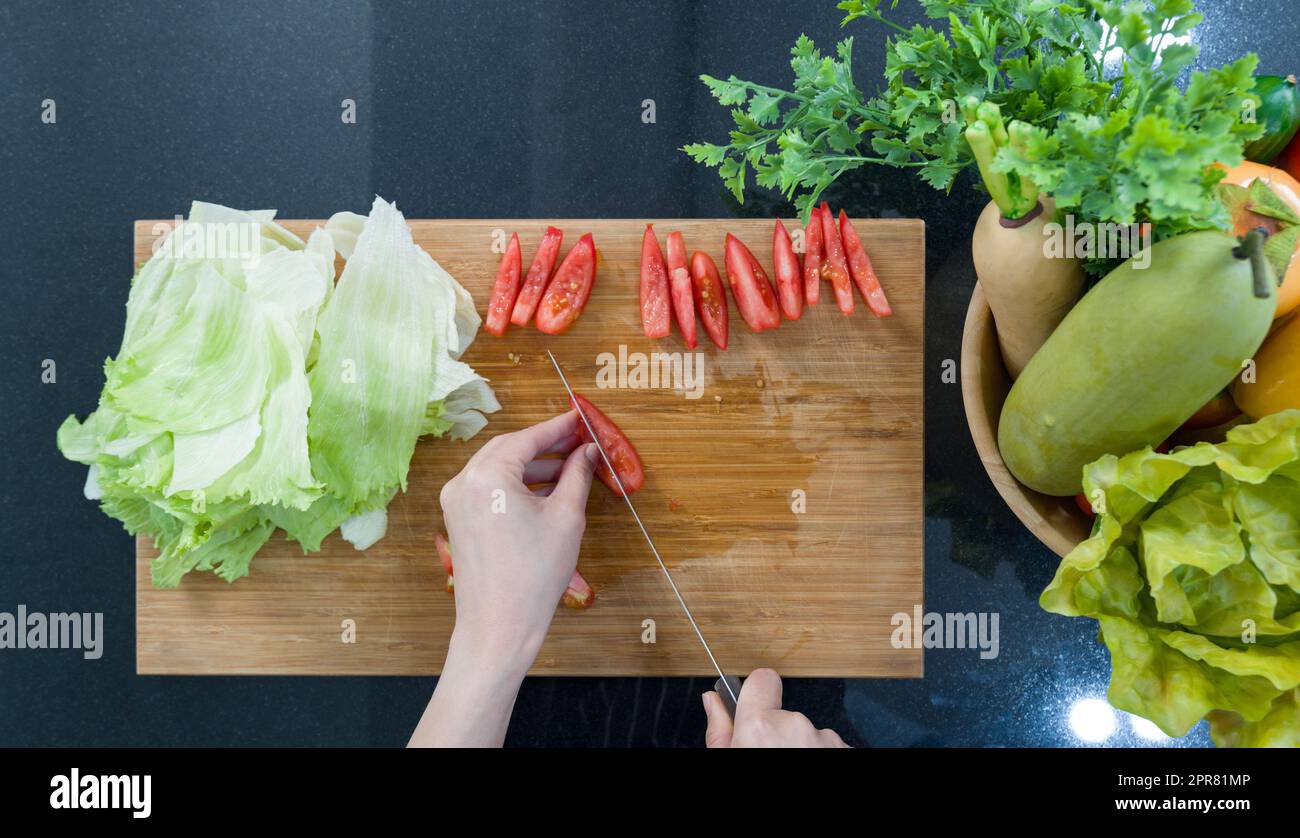 Mit der Nahaufnahme schneiden Sie rote Tomaten auf einem Holzbrett in Stücke. Draufsicht. Eine Holzschüssel mit verschiedenen Obst- und Gemüsesorten steht auf dem Küchentisch. Stockfoto