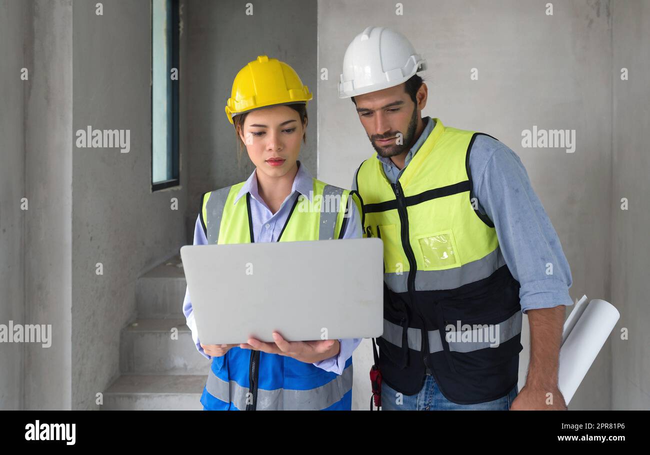 Junger Ingenieur mit Laptop-Computer erklärt dem Vorarbeiter einen Grundriss. Beide tragen einen Konstruktionshelm und eine Sicherheitsweste. Arbeitsumfeld auf der Baustelle von Wohnungsbauprojekten. Stockfoto