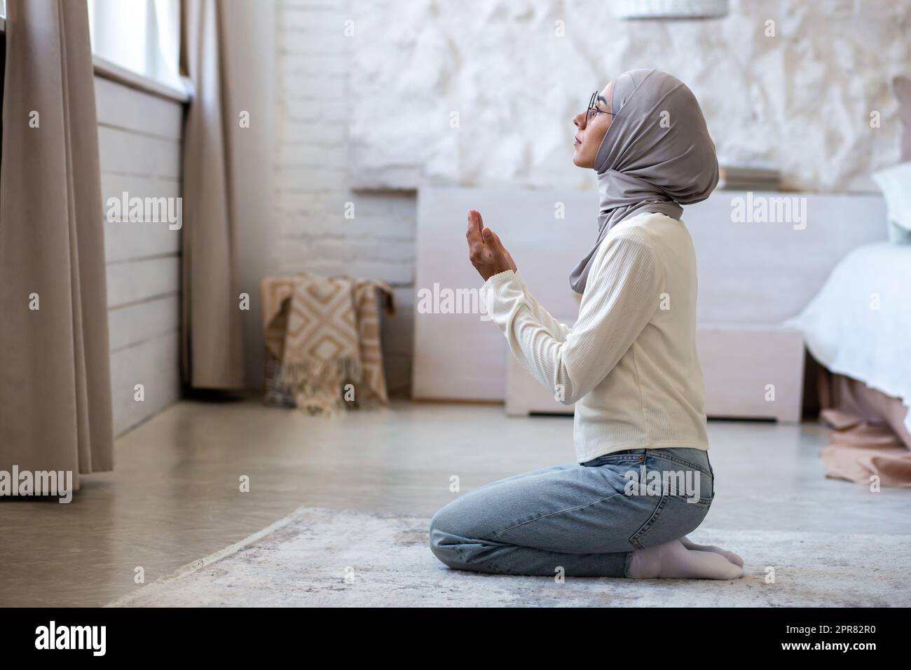 Eine junge muslimische Frau in einem Hidschab sitzt zu Hause auf ihren Knien und faltete ihre Hände, hob ihren Kopf hoch, blickt aus dem Fenster und betet. Stockfoto