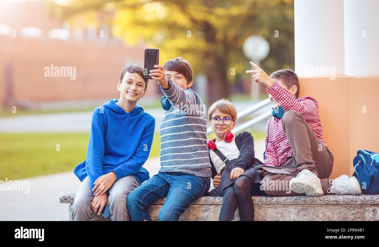 Kinder mit Rucksäcken und Kopfhörern sitzen auf der Bank im Park in der Nähe der Schule Stockfoto