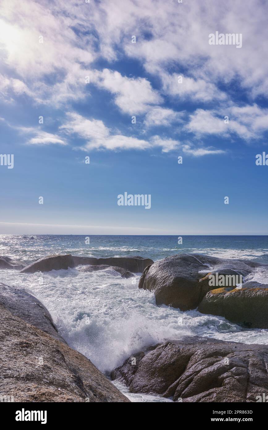 Felsen im Ozean unter blauem, wolkigen Himmel mit Kopierraum. Malerische Landschaft mit Strandwellen, die sich gegen Felsbrocken oder große Steine im Meer ergießen, an einem beliebten Sommerort in Kapstadt, Südafrika Stockfoto