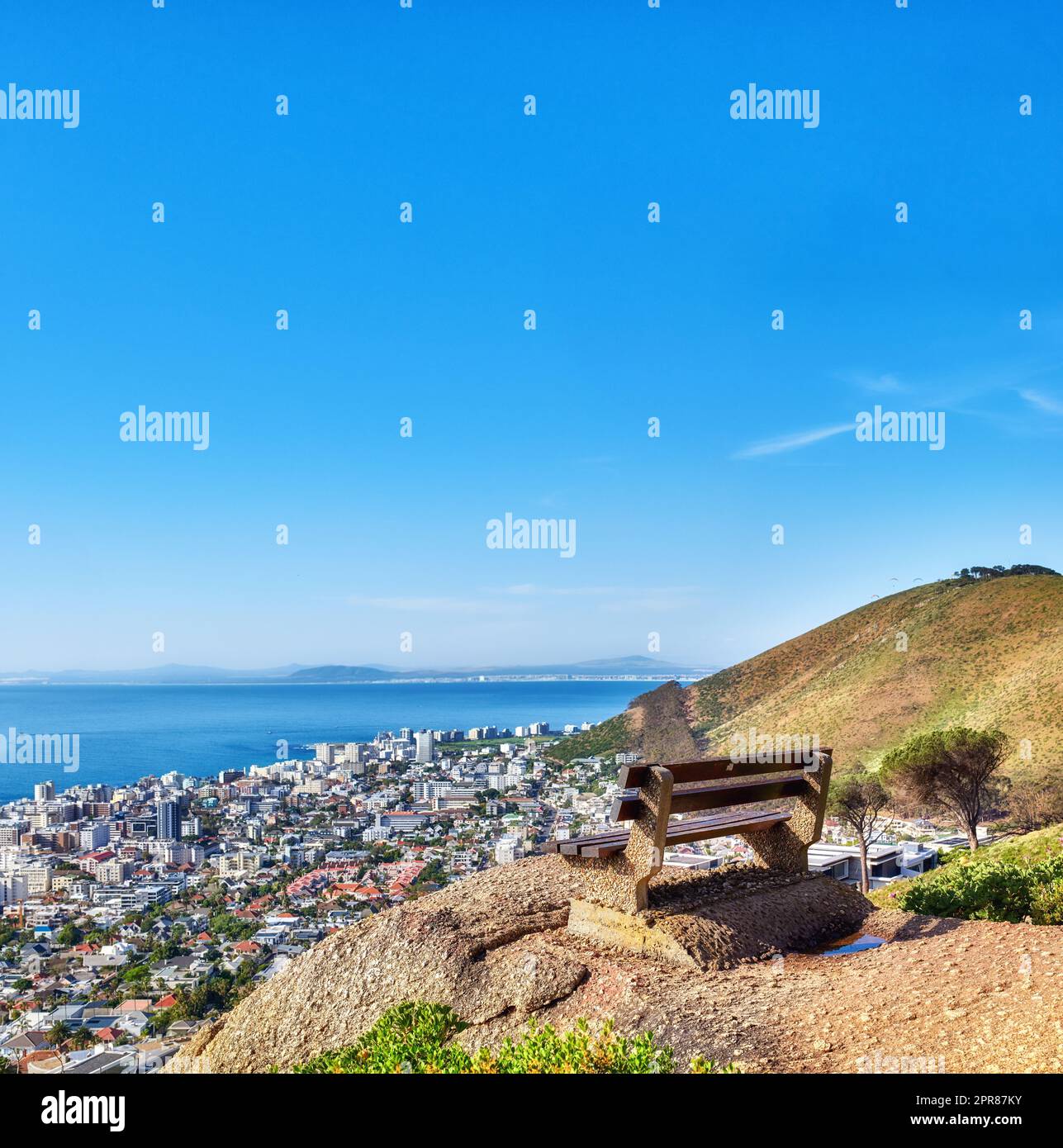 Wunderschöner Blick aus der Vogelperspektive auf eine leere Bank mit Blick auf die Stadt und das Meer in Kapstadt, Südafrika, vor dem blauen Himmel. Ein ruhiger, sonniger Tag mit einem Parkstuhl auf einer Klippe, mit malerischer Aussicht und frischer Luft Stockfoto