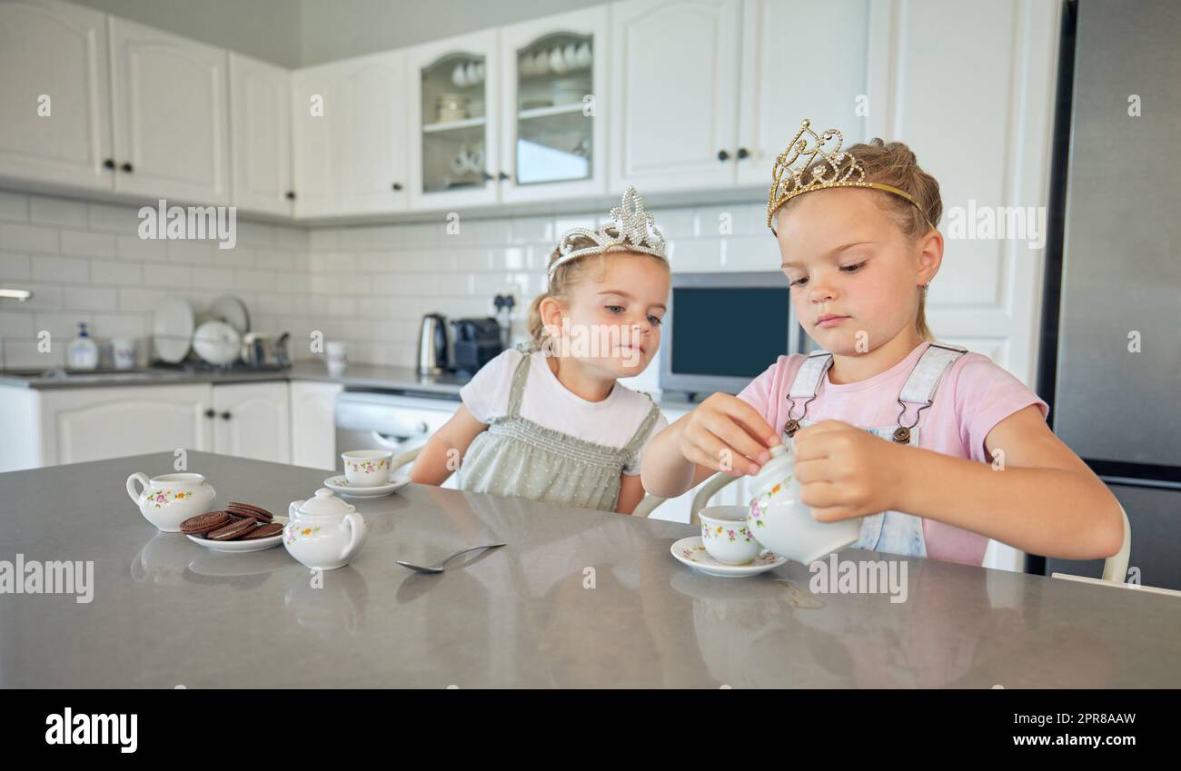 Zwei kleine Mädchen feiern zu Hause eine Teeparty. Geschwisterschwestern tragen Diademe, während sie mit dem Teeset spielen und Kekse am Küchentisch essen. Schwestern verstehen sich gut und spielen zusammen Stockfoto