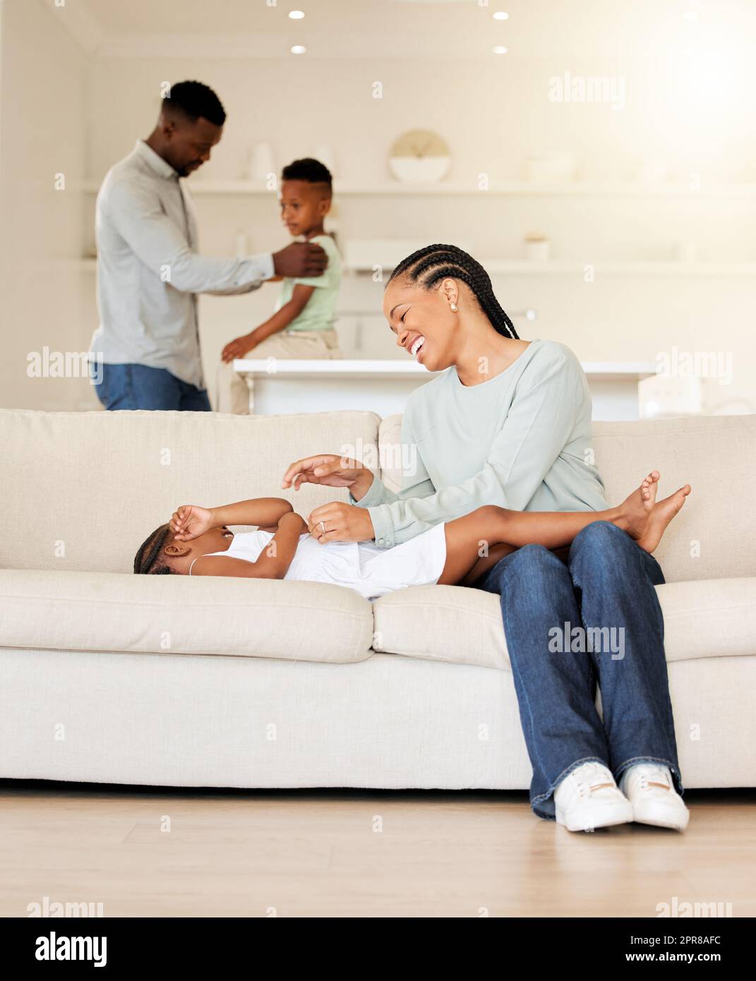 Wunderschöne junge afroamerikanische mutter, die sich mit ihrer Tochter auf dem Sofa in ihrem Wohnzimmer zu Hause anfreundet. Schwarze Frau spielt mit ihrem niedlichen kleinen Mädchen zu Hause. Dad und sein Sohn im Hintergrund Stockfoto