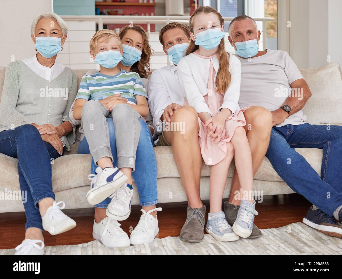 Portrait einer glücklichen Generation, weiße Familie, die Gesichtsmasken trägt, während sie sich zu Hause auf dem Sofa entspannen. Kinder, Eltern und Großeltern bleiben verantwortlich und sicher vor Krankheiten während der Covid-Pandemie Stockfoto