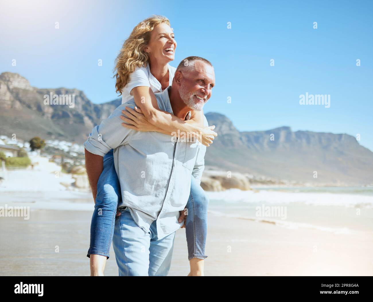 Glückliches, reifes Paar, das den Urlaub am Strand genießt. Aktiver älterer Ehemann, der seine Frau im Huckepack mitnimmt und dabei einen sonnigen Tag im Freien genießt. Energiegeladener Mann und Frau, die im Urlaub Spaß haben Stockfoto