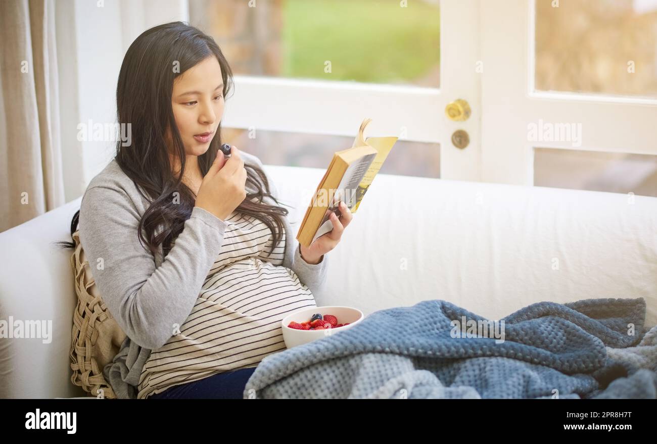 Gesund zu essen wurde gerade doppelt so wichtig. Eine attraktive junge Schwangere, die Obst isst und ein Buch liest, während sie sich zu Hause auf dem Sofa entspannt. Stockfoto