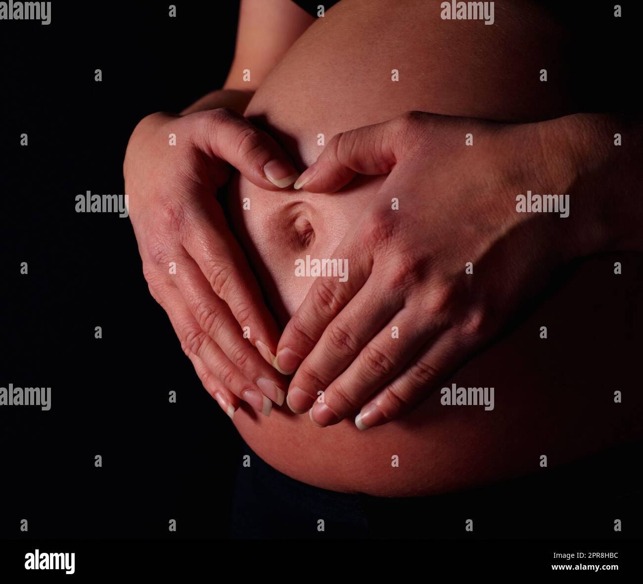 Schwanger Frau macht Herzform mit Händen gegen schwarz. Detailaufnahme einer Schwangeren, die mit Händen vor schwarzem Hintergrund Herzform formt. Stockfoto