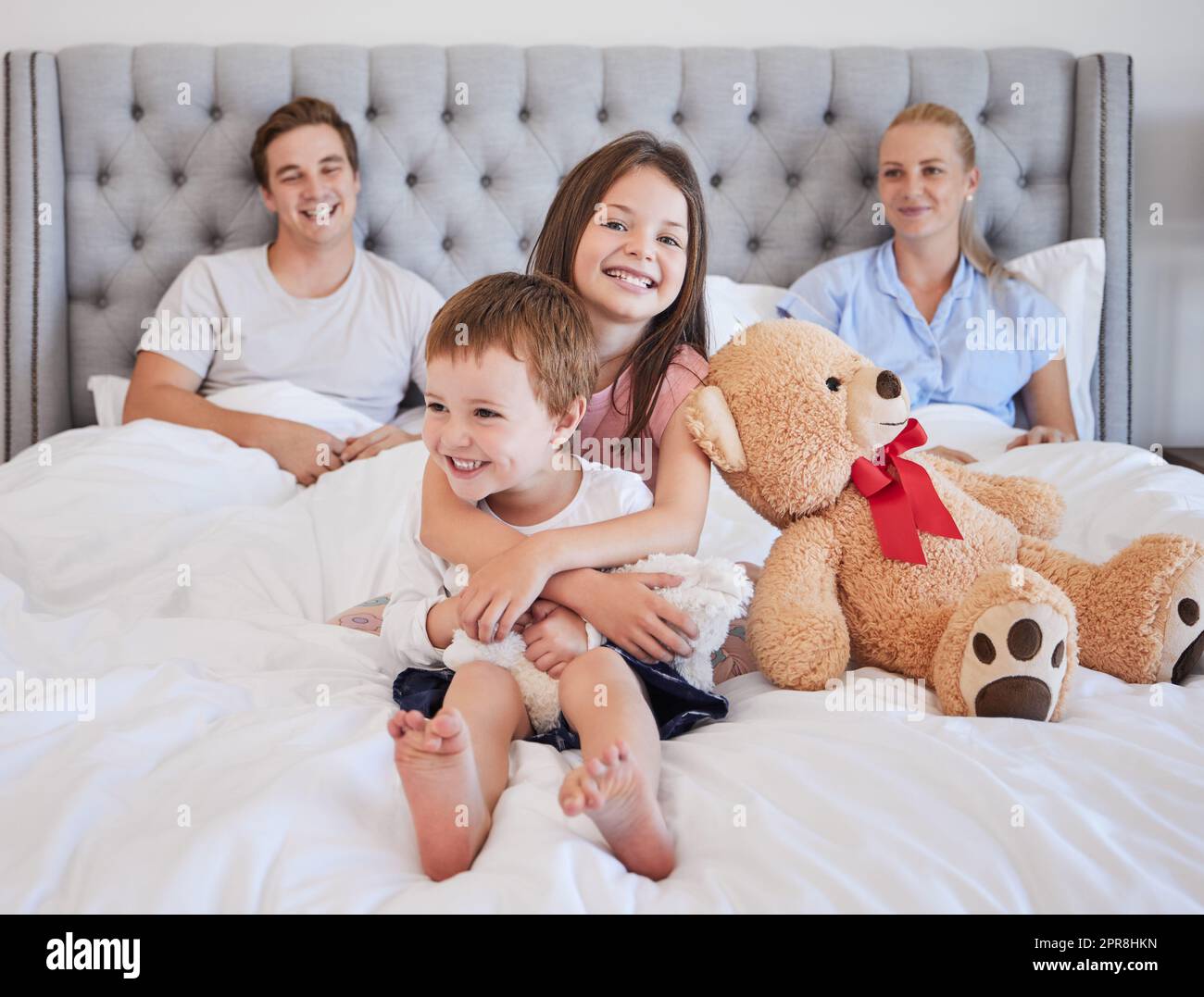 Ein bezauberndes kleines Mädchen umarmt ihren jüngeren Bruder mit ihrem Teddybär neben ihr, während sie mit ihren Eltern im Hintergrund auf einem Bett sitzt. Ich liebe es, dass sich die weiße Familie anfreundet und am Morgen Zeit miteinander verbringt. Eine große Schwester zu haben, bedeutet, dass du eine hast Stockfoto