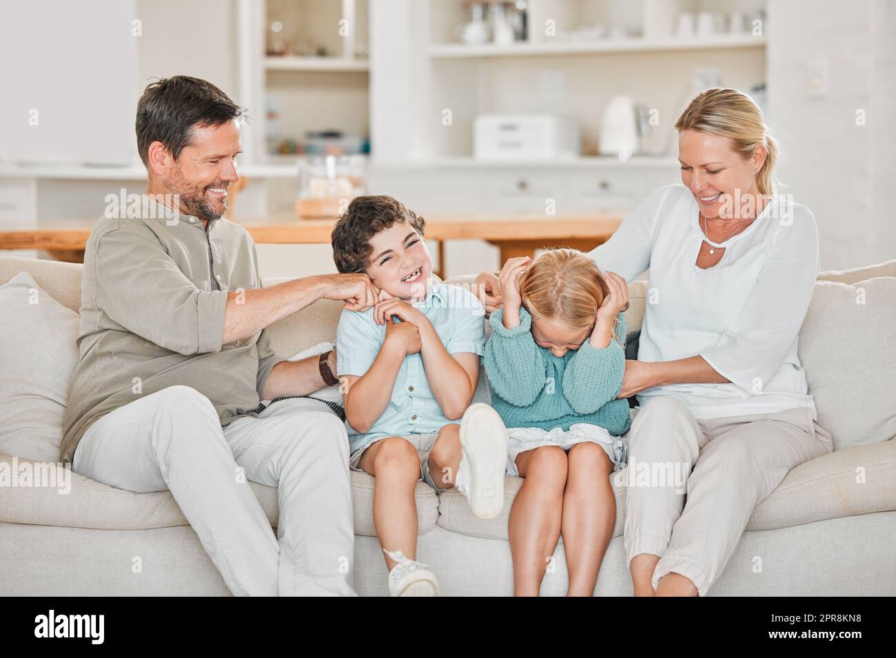 Kitzeln, kitzeln. Ein entzückender kleiner Junge und ein Mädchen, das von ihren Eltern auf dem Sofa zu Hause gekitzelt wird. Stockfoto