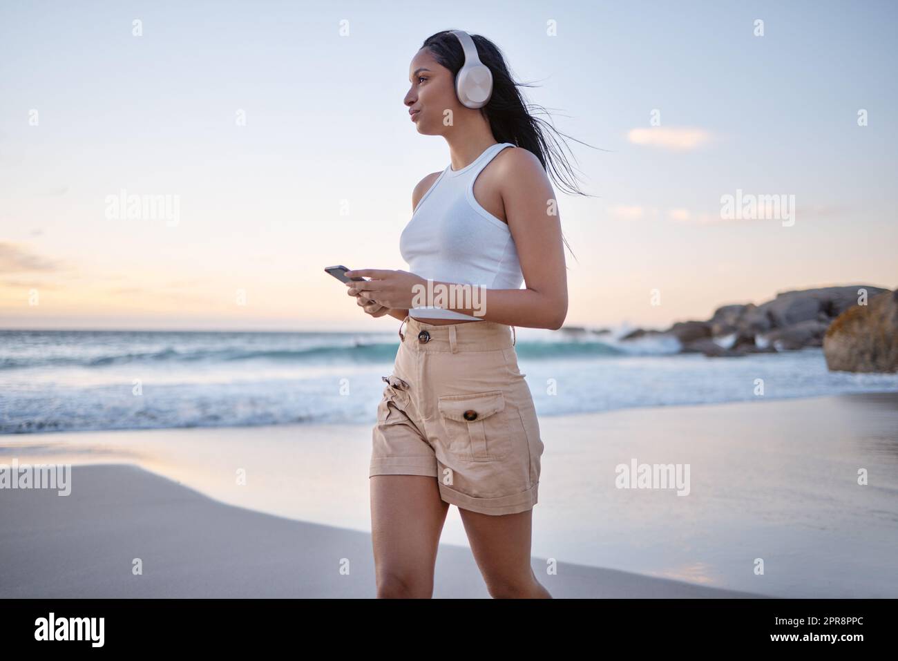 Nur ich, Musik und das Meer. Eine Frau, die Kopfhörer trägt und ihr Handy benutzt, während sie am Strand spazieren geht. Stockfoto